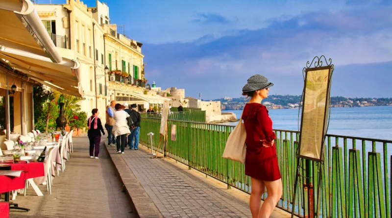 Woman reads menu by the sea with buildings.