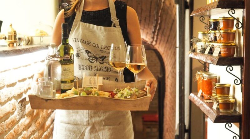 A waitress in a vintage apron serves wine and appetizers in a cozy, rustic indoor setting.
