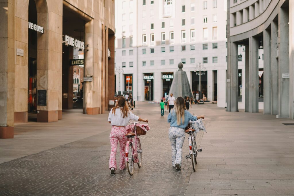 deux jeunes femmes à vélo dans une piazza italienne, vêtues de façon décontractée