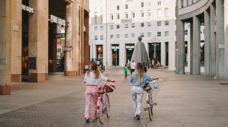 deux jeunes femmes à vélo dans une piazza italienne, vêtues de façon décontractée